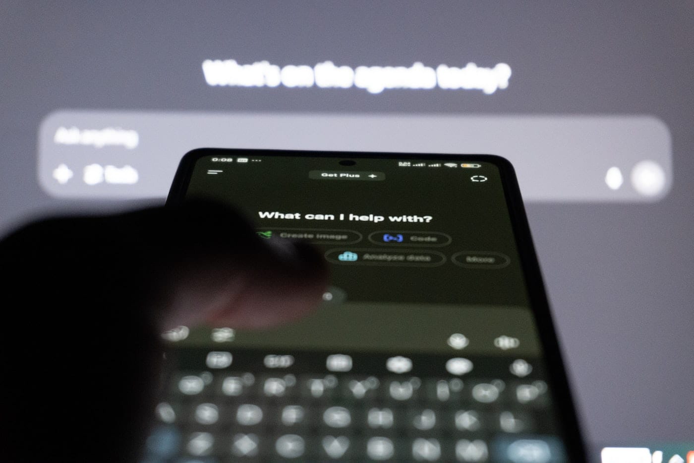 Pessoa segurando smartphone com tela de chatbot de IA aberta perguntando 'What can I help with?', tela de fundo desfocada mostrando interface de assistente virtual