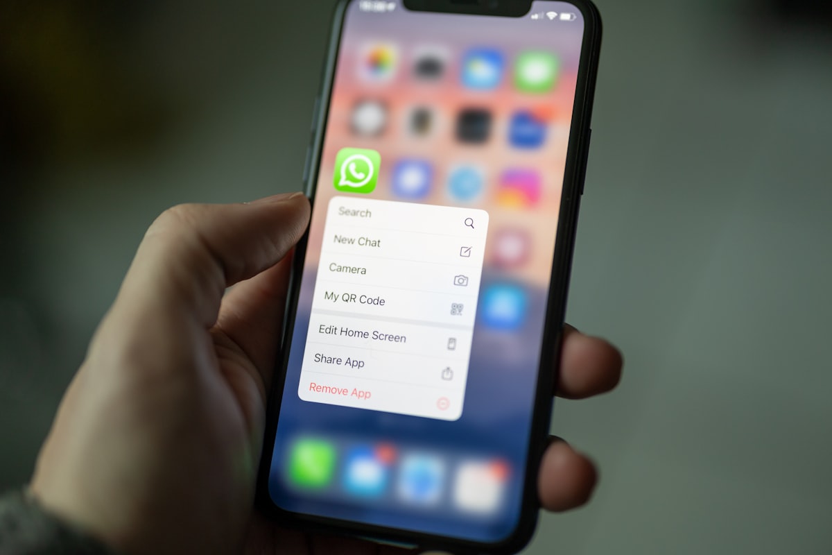 iPhone segurado na mão mostrando o ícone do WhatsApp em destaque com menu de contexto aberto (Search, New Chat, Camera, My QR Code) — onde começa o atalho pra criar lista de transmissão